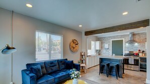 Een moderne woonkamer met een blauwe bank, een gele salontafel, en een keuken met een betegelde achterwand en roestvrijstalen apparatuur.