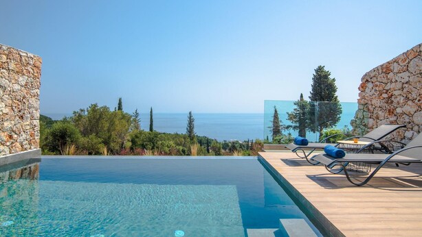 Ein klares, blaues Schwimmbecken mit Holzterrasse und Liegestühlen, mit Blick auf eine schöne Landschaft aus Meer und Grün.