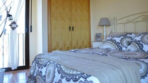 A bedroom with a large wooden wardrobe, a bed with patterned bedding, a nightstand with a lamp, and a window with curtains.