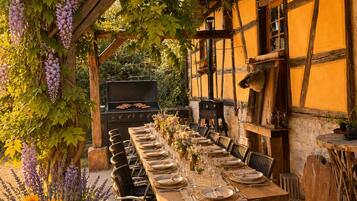An outdoor dining area with a long wooden table set for a meal, surrounded by wicker chairs, under a pergola with hanging wisteria.