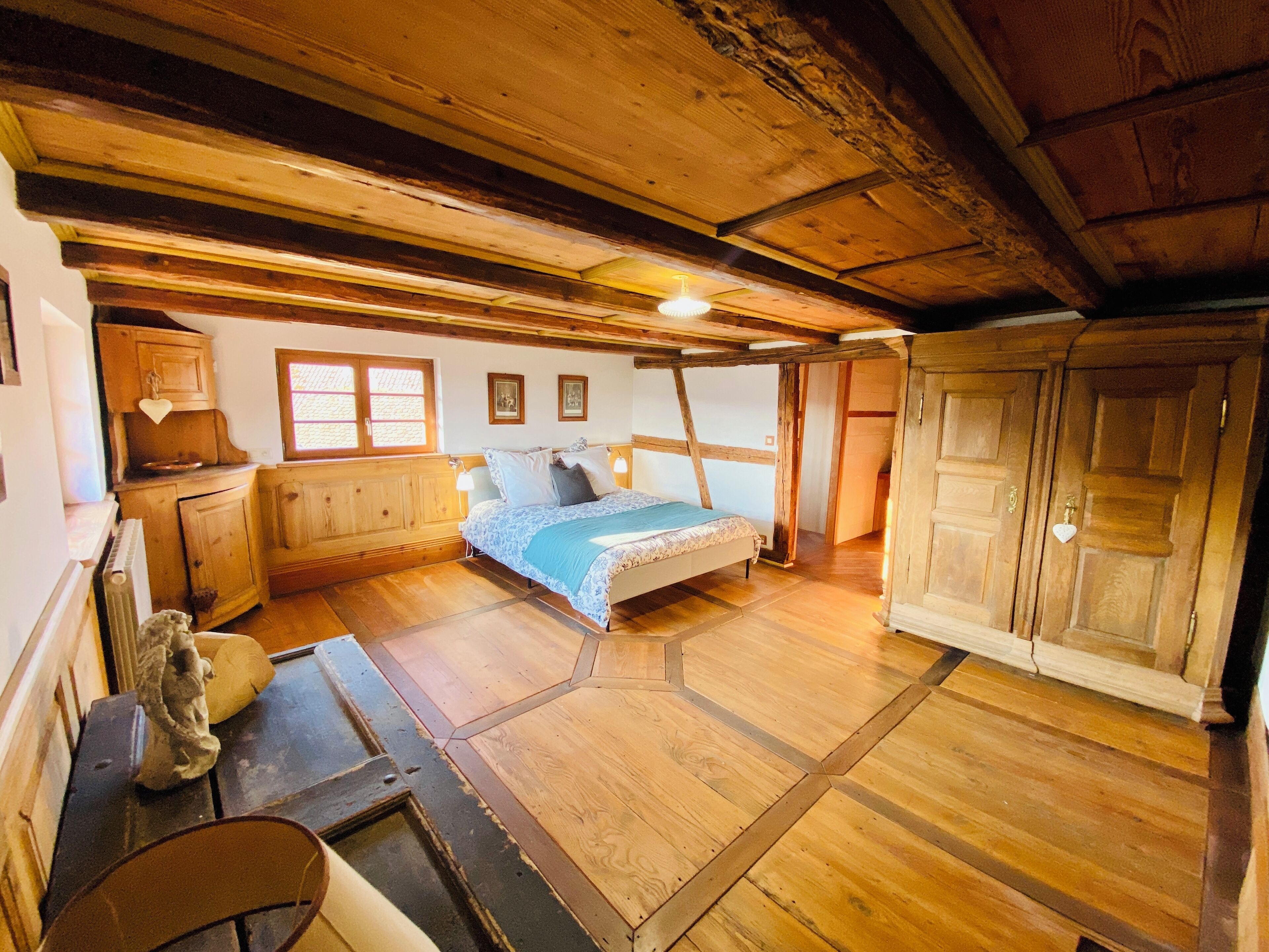 Een rustieke slaapkamer met een houten plafond, een bed met een blauwe deken, houten kasten en een klein beeldje op een tafel.