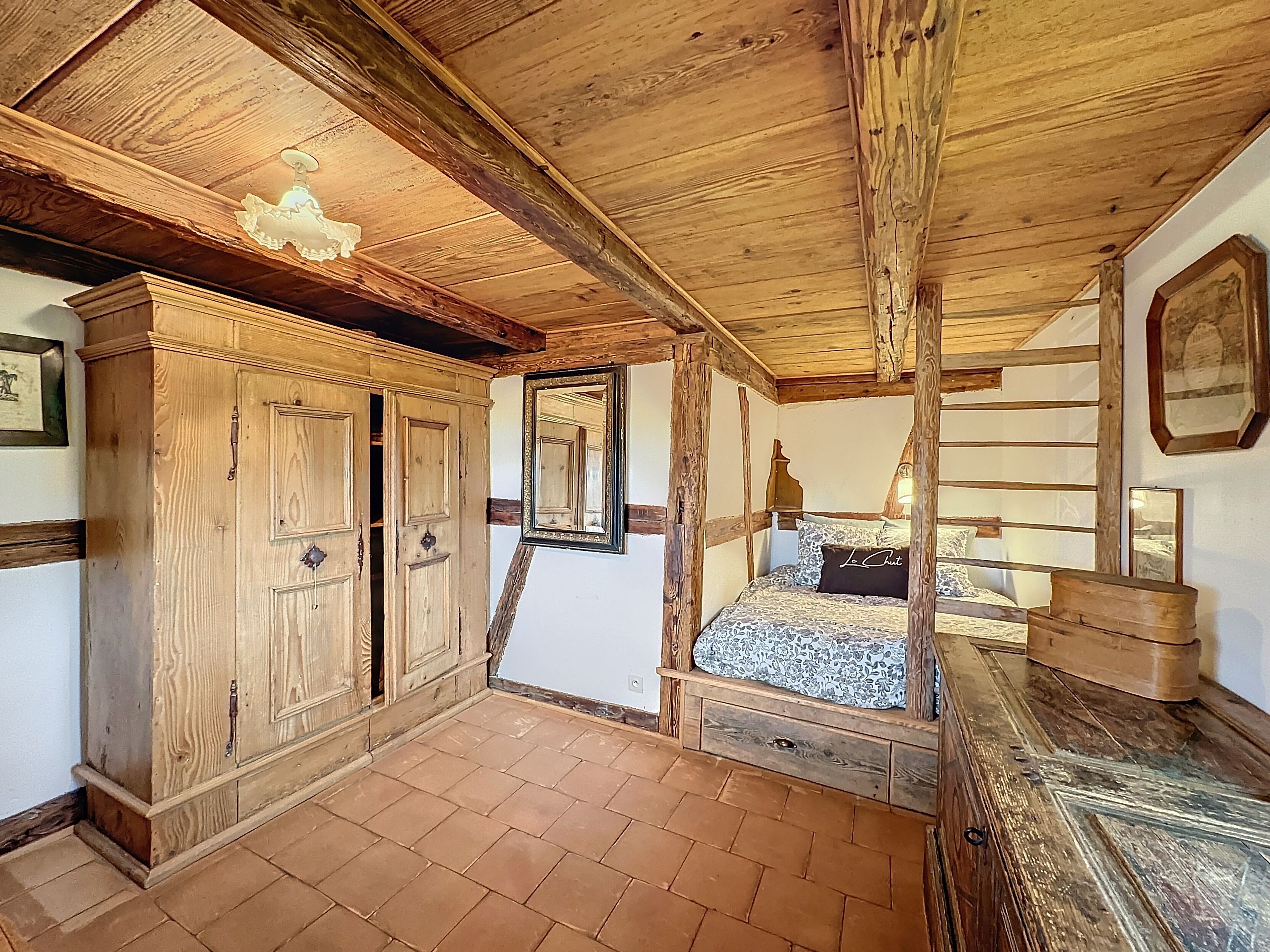 Une chambre à coucher au style rustique, avec des poutres apparentes, une grande armoire, un lit recouvert d’une literie à motifs et un miroir accroché au mur.