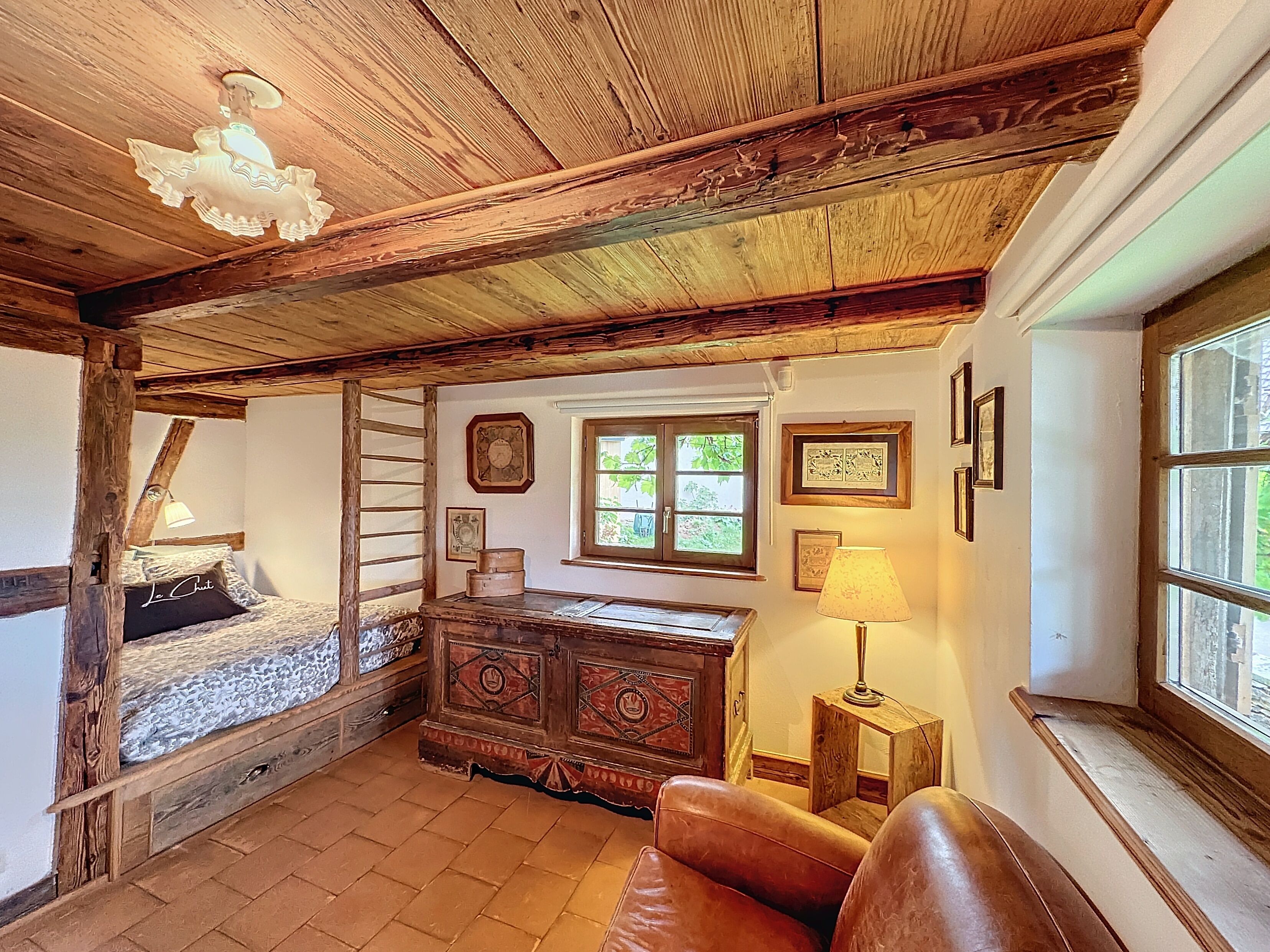 Quarto rústico com teto de madeira, cama com cabeceira, cômoda, abajur e janela com vista para o verde.