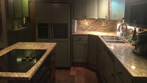 Une cuisine dotée de plans de travail en granit, d’une cuisinière encastrée et de meubles de couleur verte. On y trouve également un tableau noir sur lequel est écrit un message de bienvenue.