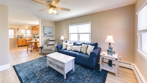 A living room with a blue sofa, a coffee table, a lamp, and a ceiling fan. - Cozy Villas Cottage w/ Deck, 1/2 Block to Beach! (Villas)