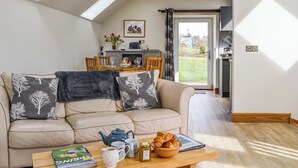 A cozy living room with a sofa, a coffee table with books and pastries, and a view of a dining area and garden through a glass door. - 2 bedroom accommodation in Fettercairn (Fettercairn)