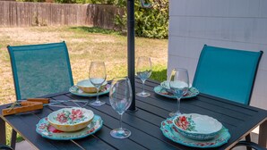 Una mesa de comedor dispuesta con platos estampados de flores y copas de vino, rodeada de sillas color turquesa, en un entorno al aire libre.