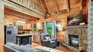Ein gemütliches Blockhaus mit Kamin aus Stein, Holzwänden, einer Küchenzeile und einer Sitzecke mit Sofa und Sesseln.