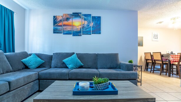 Ein modernes Wohnzimmer mit einem grauen Ecksofa, einem Couchtisch mit Kerze und Pflanze sowie einem Essbereich mit Tisch und Stühlen.