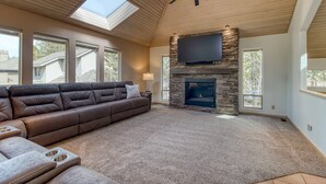 A spacious living room with a large sectional sofa, a stone fireplace, and a flat-screen TV mounted on the wall. - Modern Sunriver Luxury Home with Fenced Turf Pet Yard, Large Kids Room, SHARC !! (Sunriver)