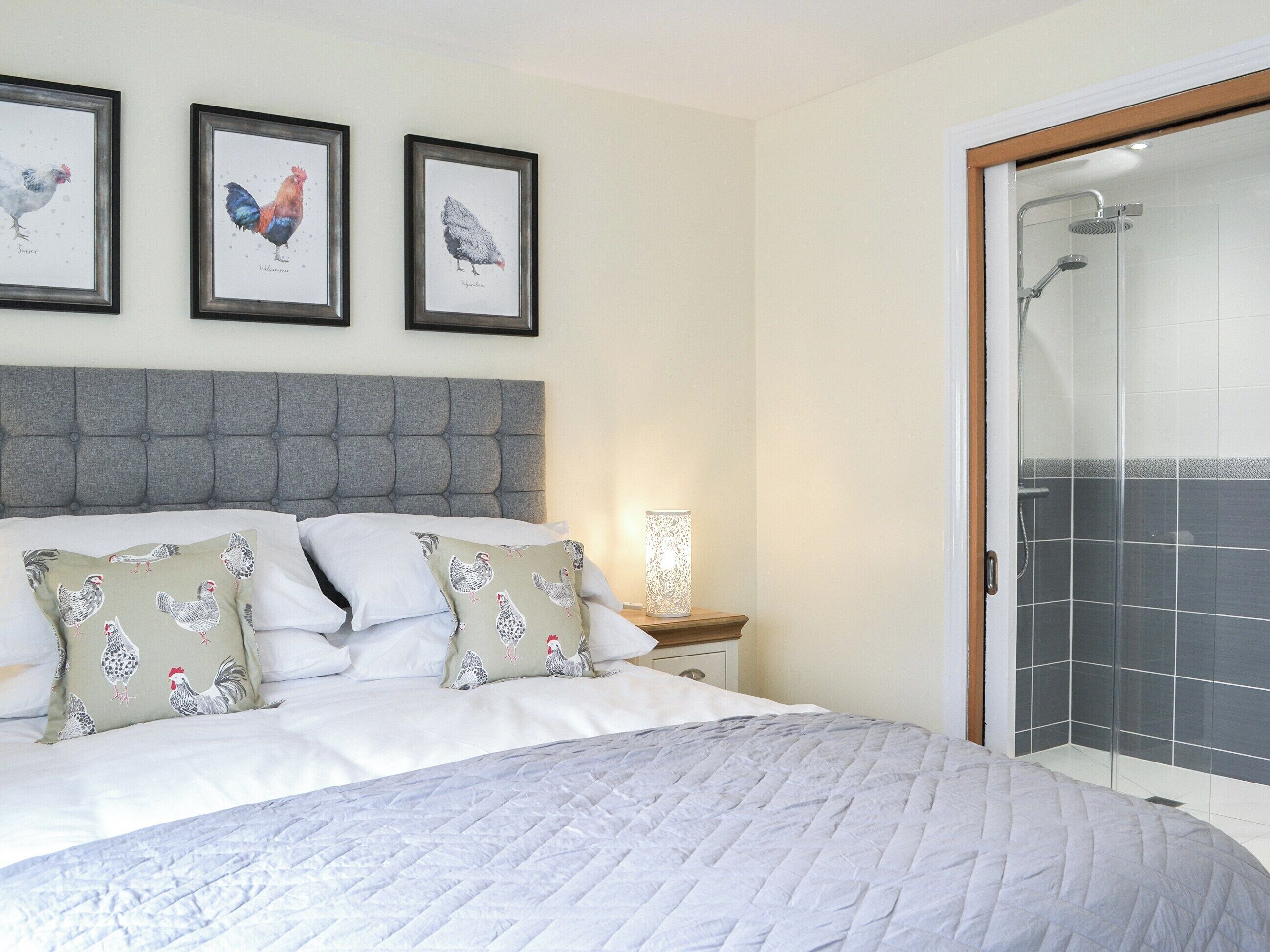 Une chambre avec une tête de lit capitonnée grise, un lit avec une literie blanche et des coussins à motifs de poulets, une table de chevet avec une lampe et une illustration encadrée de poulets accrochée au mur.