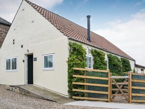 Een gelijkvloers huis met een witte gevel, een rode dakbedekking en een schoorsteen. Voor is een houten poort en het huis wordt omringd door grind.