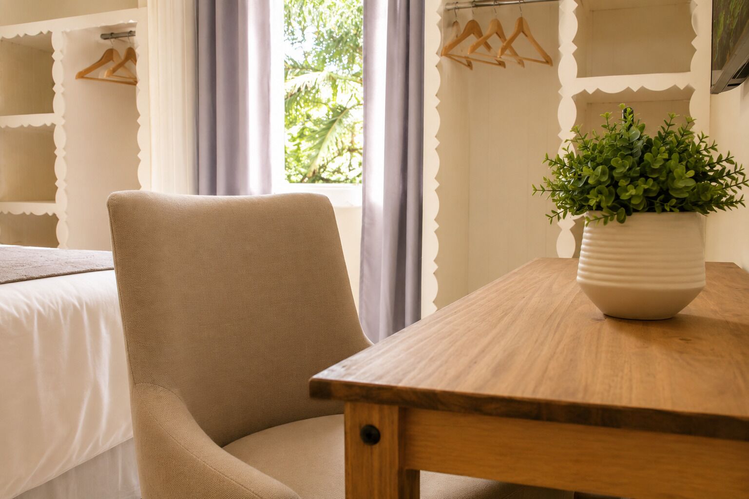 A room with a bed, a chair, a wooden table, a potted plant, and a window with curtains.