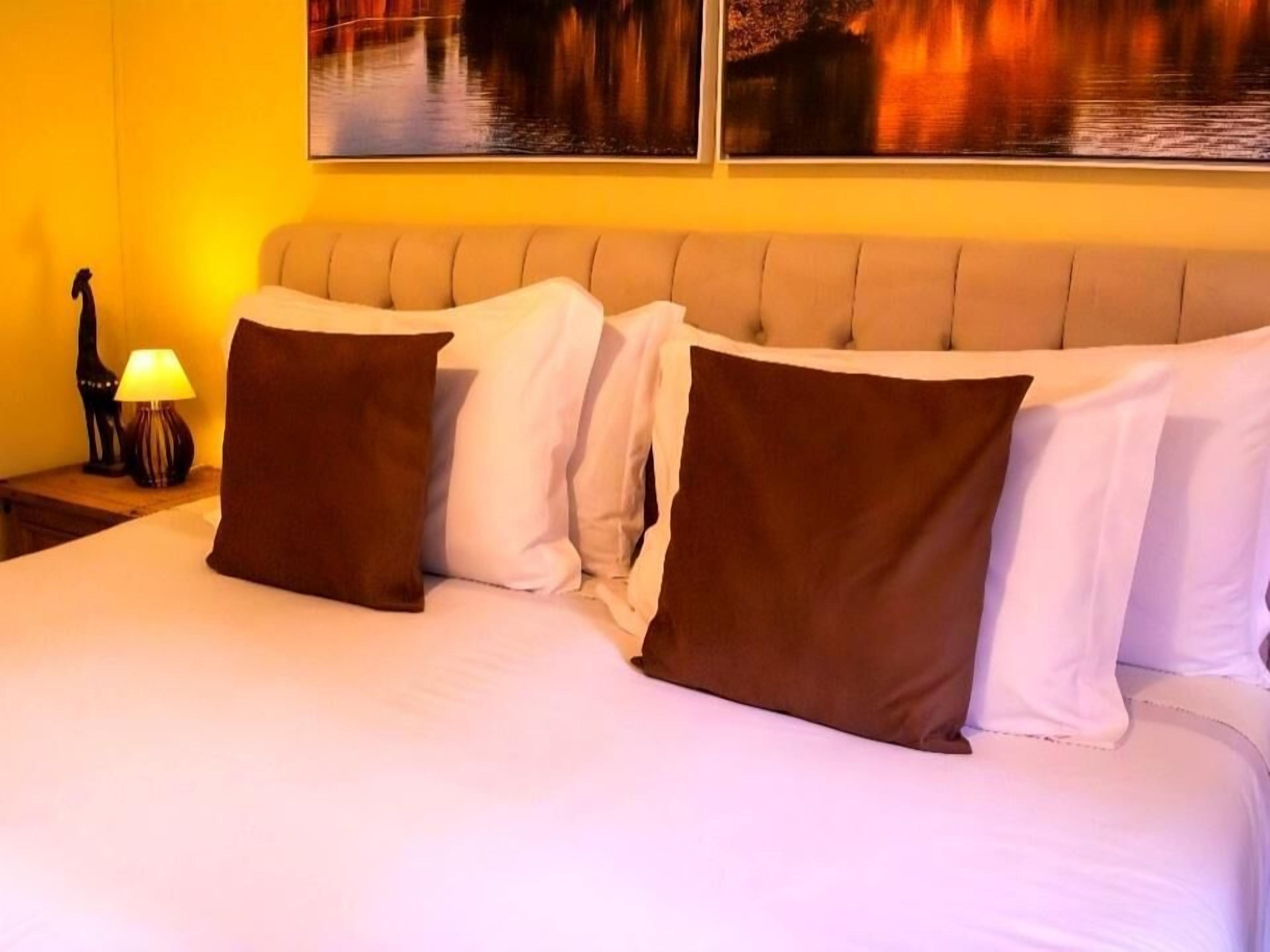 A hotel room with a bed, pillows, a nightstand with a lamp, and a painting on the wall.