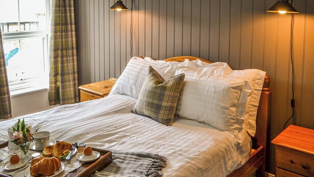Una cama bien hecha con sábanas blancas, una almohada de cuadros, y una bandeja con cruasanes y una taza de café. Hay dos lámparas de noche y una mesita de madera junto a la cama.