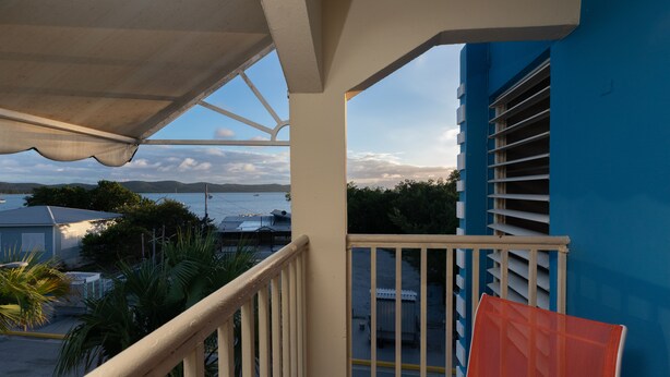 A balcony with a view of the sea and mountains, a white railing, and a blue building in the background. - Red Sunset Beach Club (Cabo Rojo)