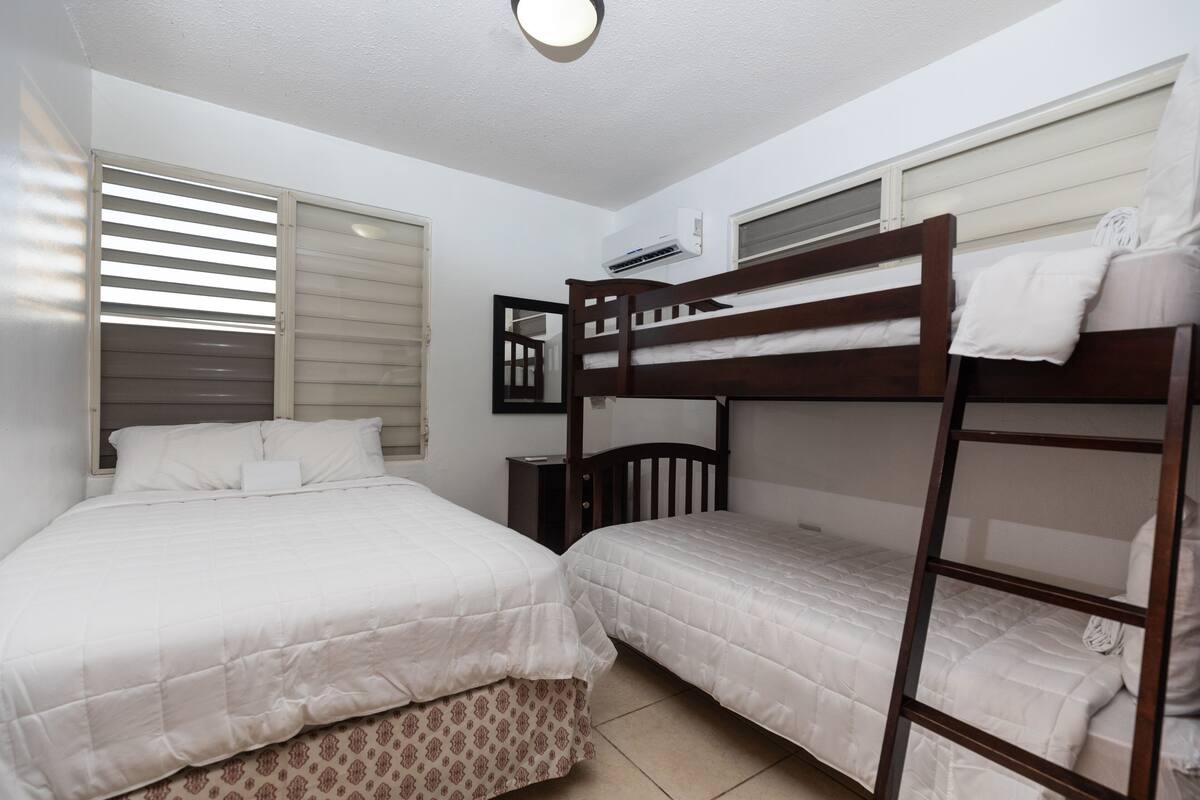 A room with two single beds, a bunk bed, a window with blinds, and an air conditioning unit.