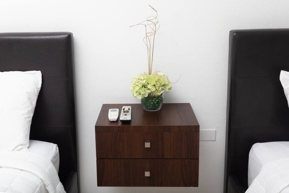 A hotel room with two black leather headboards, a wooden nightstand with a vase of green flowers, and a remote control.