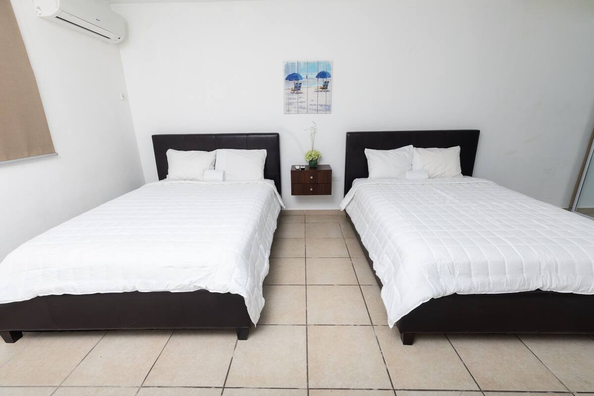 A hotel room with two single beds, a small nightstand with a vase, and a beach-themed wall art.