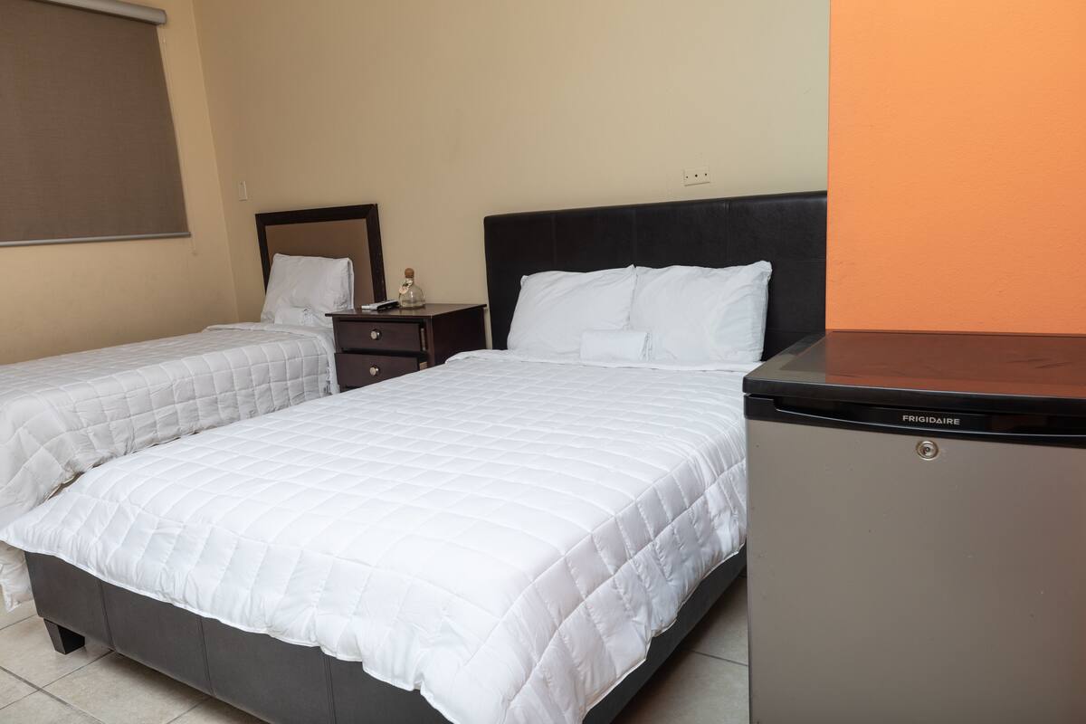 A hotel room with two beds, a small table, a nightstand, and a refrigerator.