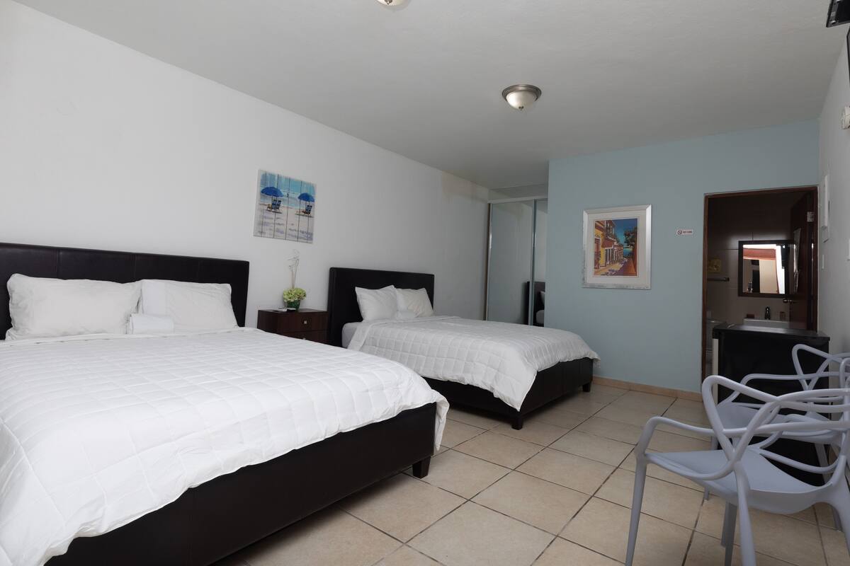 A hotel room with two beds, a small table, a chair, and a bathroom visible through an open door.