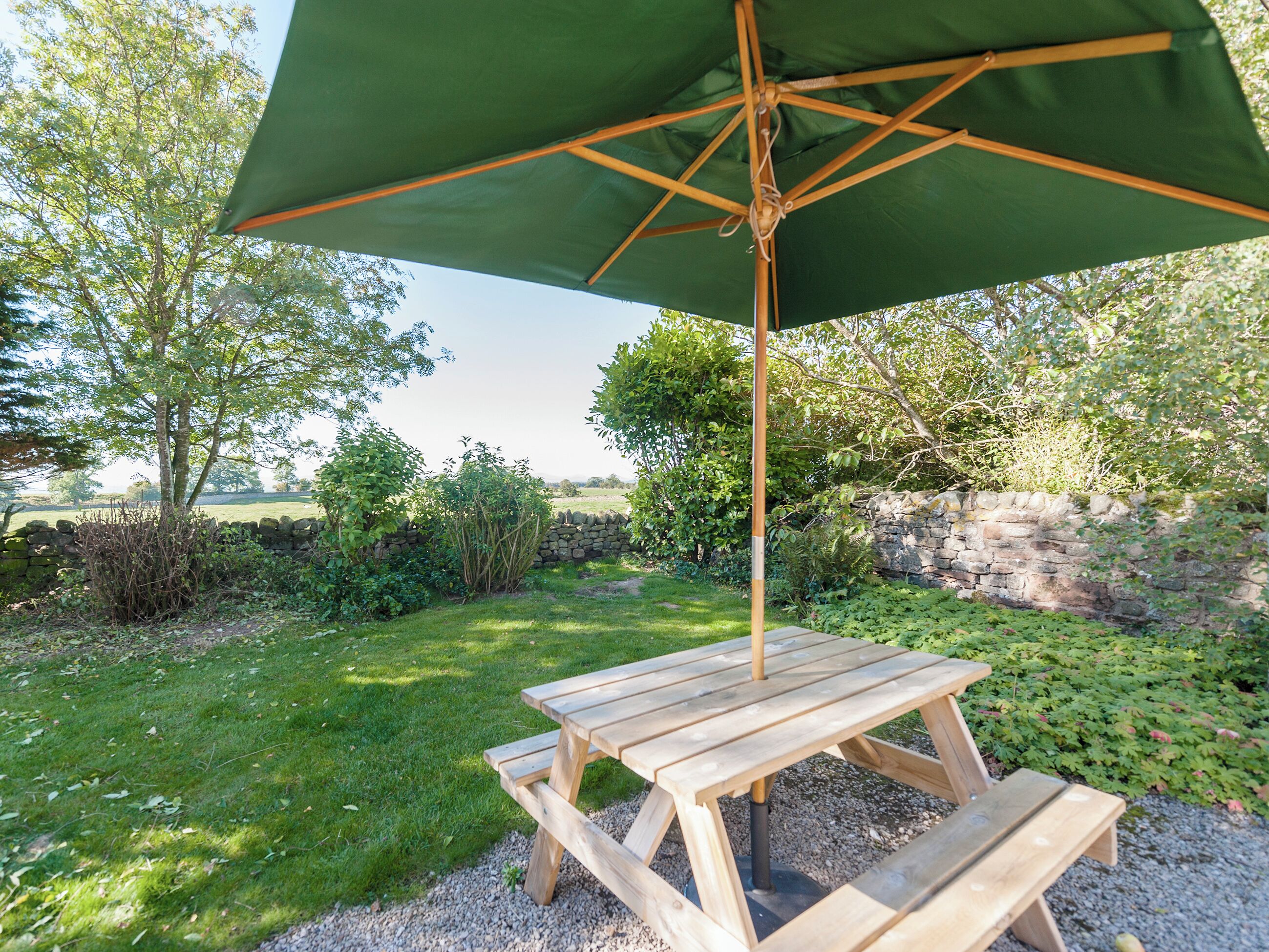 A wooden picnic table with a green umbrella, surrounded by a stone wall and greenery.