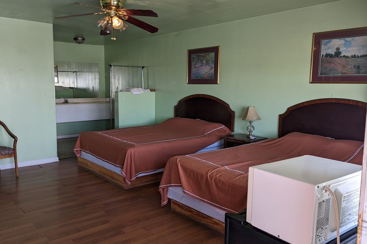 A room with two beds, a chair, a desk, a ceiling fan, and a painting on the wall.