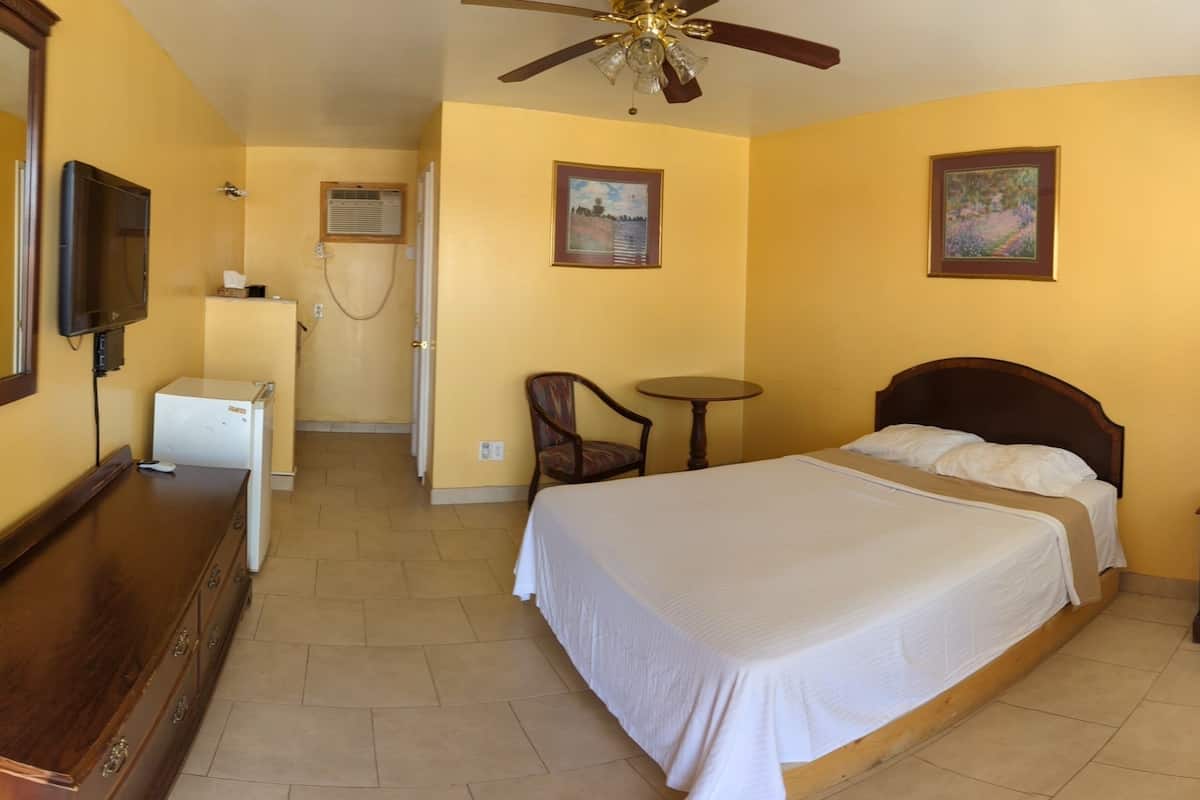A hotel room with a bed, a desk, a chair, a TV, a ceiling fan, and artwork on the walls.