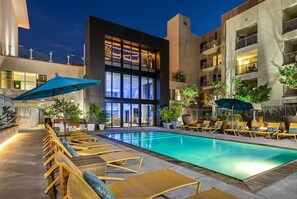 Un hotel moderno con piscina, sillones de descanso y un edificio grande con balcones.