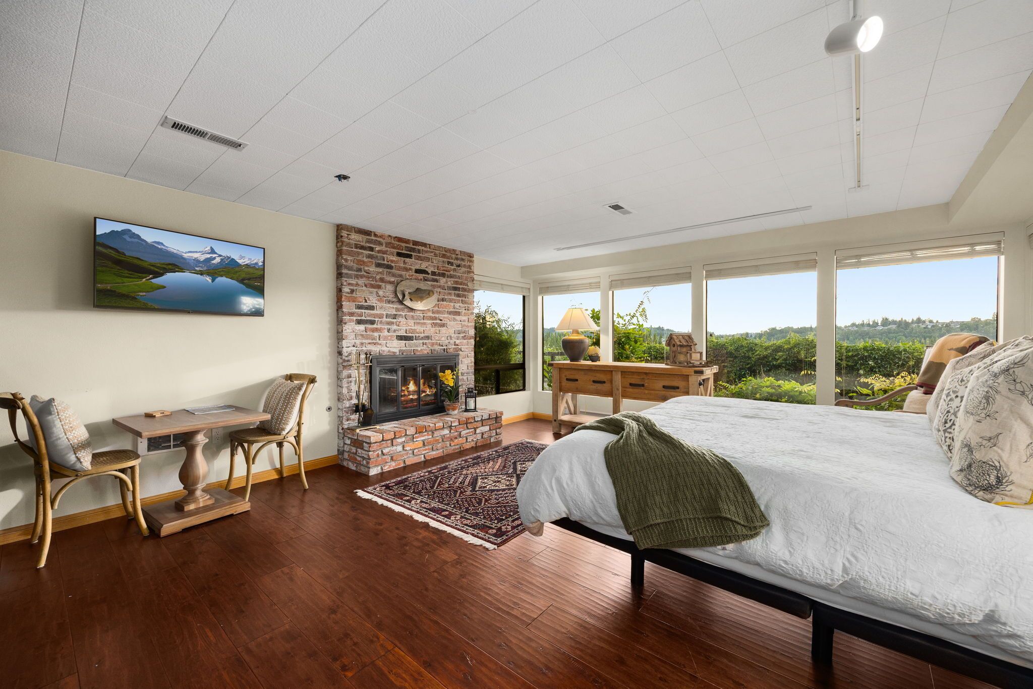 A bedroom with a fireplace, a bed, a desk, and a large window with a view.