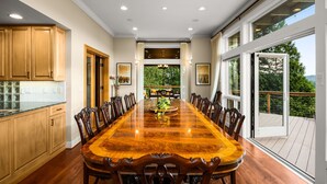 A spacious dining room with a large wooden table and chairs, wooden cabinets, and large windows offering a view of greenery.