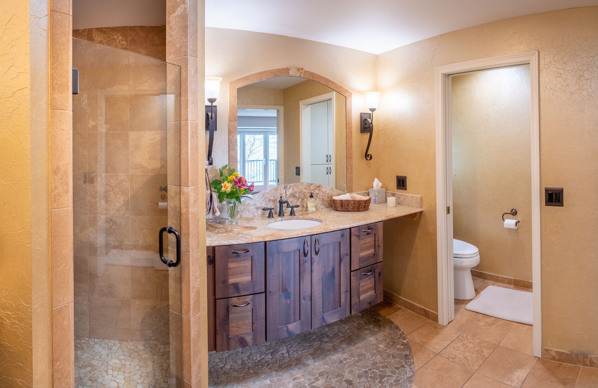 A bathroom with a glass-enclosed shower, a vanity with a marble countertop, wooden cabinets, and a large mirror.