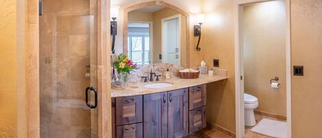 A bathroom with a glass-enclosed shower, a vanity with a marble countertop, wooden cabinets, and a large mirror.