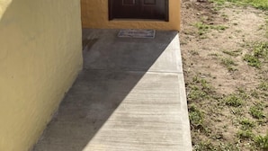 Un bâtiment jaune avec une porte brun foncé, un petit porche en béton et une cour couverte d’herbe.