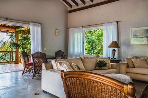 A living room with a sofa, chairs, a table, and a lamp. There are windows with curtains, a view of greenery outside, and a ceiling with wooden beams. - Mansão Sobreomar - Ilhabela (Ilhabela)