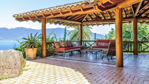 A covered outdoor seating area with a wooden pergola, red cushioned sofas, and a tiled floor. - Mansão Sobreomar - Ilhabela (Ilhabela)