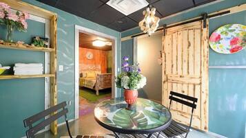 A room with a round glass table, two chairs, a wooden bench, a potted plant, a wooden door, and a wall-mounted shelf with flowers.