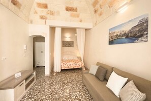 A bedroom with a stone wall, a bed with a floral bedspread, a sofa with pillows, a painting on the wall, and a TV on a wooden stand.