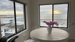 A room with a round table, clear chairs, a white vase with flowers, and large windows overlooking a beach and ocean. - Beachfront Penthouse at The Entrance - Beach Pool right infront (The Entrance)