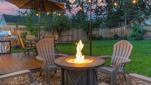 Un patio al aire libre con una mesa redonda, dos sillas Adirondack, un brasero y luces colgantes.