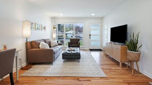 Una sala de estar moderna con un sofá, reposapiés, televisor y plantas en macetas.