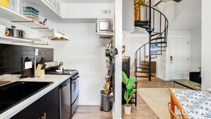 Une cuisine moderne avec des meubles noirs, un évier en acier inoxydable et un four encastré. On y trouve une petite télévision posée sur une étagère blanche et un escalier en colimaçon menant à un autre niveau.