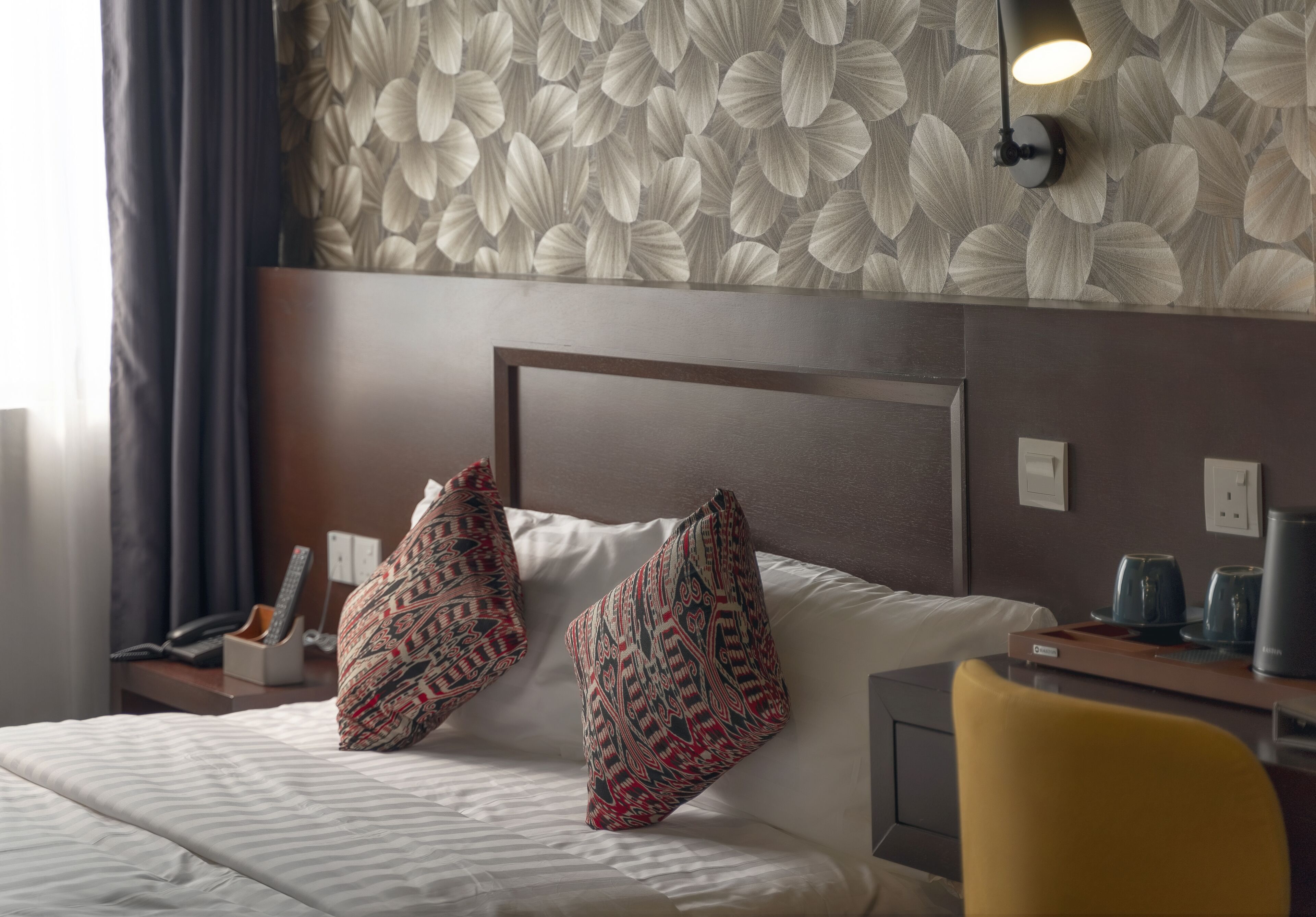 Habitación de hotel con cama, dos almohadas estampadas, mesita de noche con lámpara, una silla y pared con diseño de hojas.