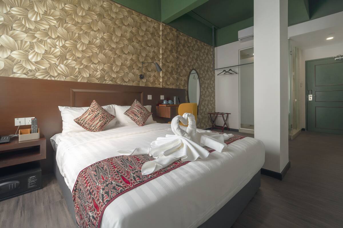 A hotel room with a large bed, a headboard, a nightstand, a bedside table, a wall-mounted mirror, a wardrobe, and a door.