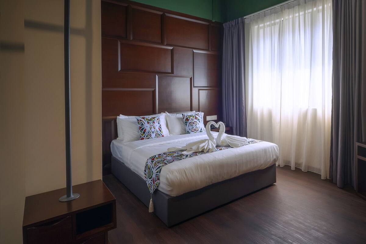 A hotel room with a large bed, a wooden headboard, a bedside table, and a window with curtains.