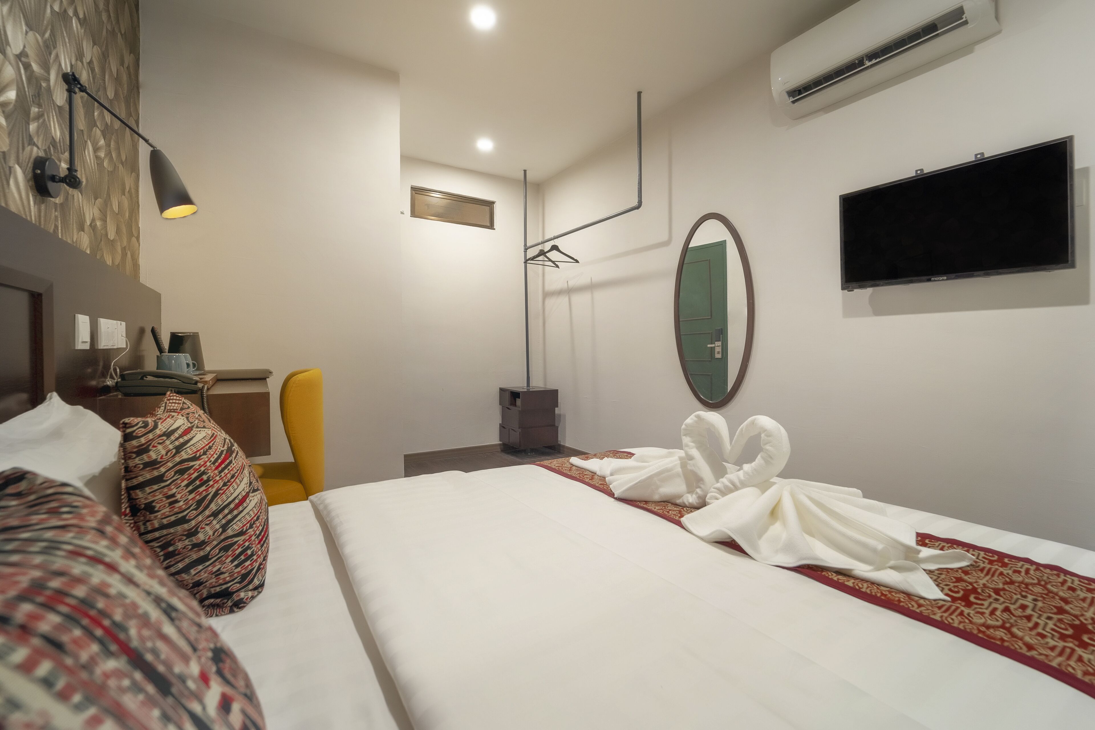 Una habitación de hotel con una cama bien hecha, un televisor de pantalla plana montado en la pared, un espejo y una mesita de noche con una lámpara.