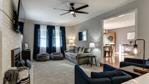A modern living room with a fireplace, a ceiling fan, and a dining area with a wooden table and chairs. - Updated home, hot tub, fire pit close to Asheville (Candler)