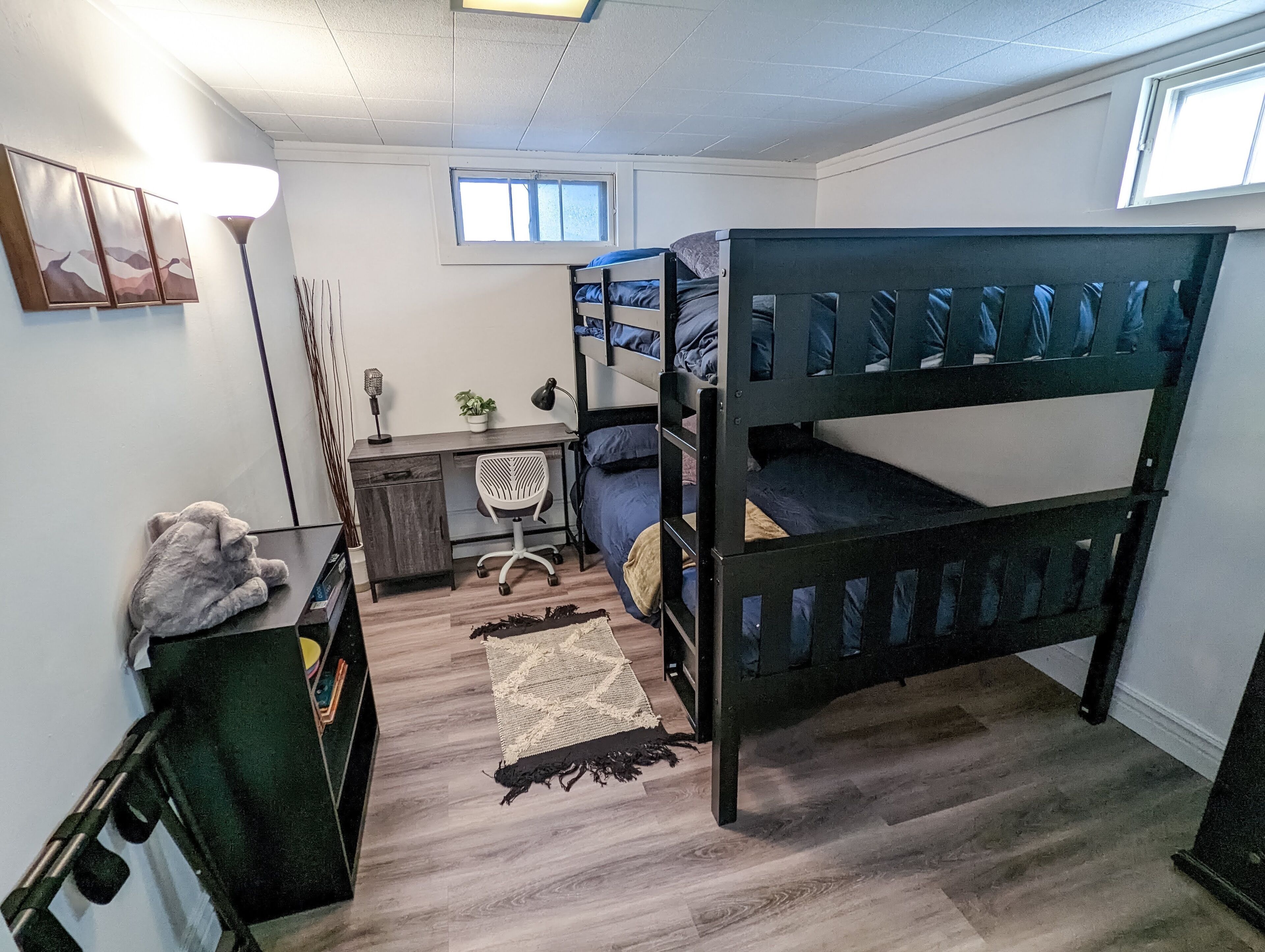 A dormitory room with bunk beds, a desk, a chair, a lamp, a plant, and a window.