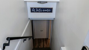 Uma escadaria com corrimão de madeira, um sinal com a palavra 'hi' e um aviso de 'Welcome' acima de uma porta.