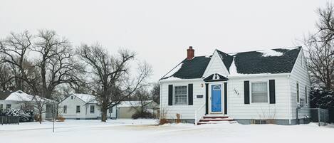 Een wit huis met een blauwe deur, omgeven door een met sneeuw bedekte grond en kale bomen.